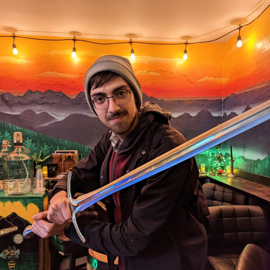 A tall, dark-haired man with rectangular glasses wearing a light blue beanie, red hoodie, and black jacket, holding a medieval sword in a defensive position
